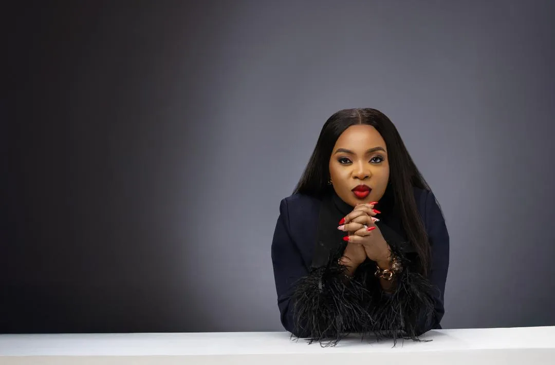 Mabel Adeteye leaning forward in a dark feathered jacket against a charcoal backdrop