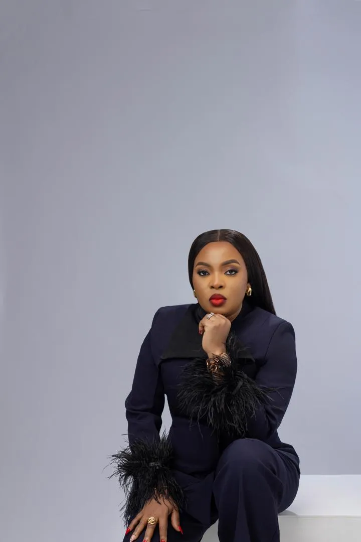 Mabel Adeteye seated in a navy suit with feathered cuffs against a pale grey backdrop